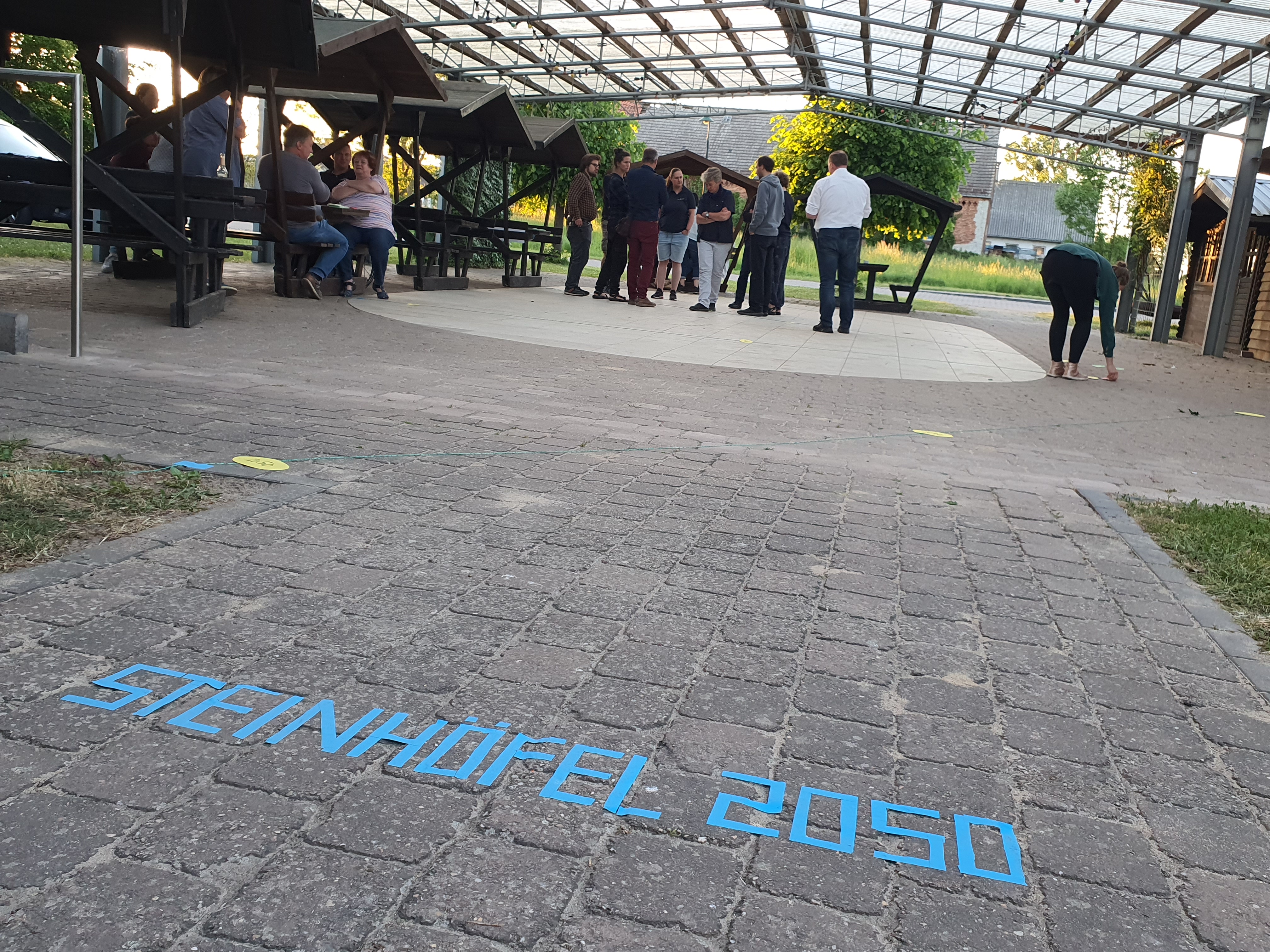 Im Vordergrund ist der Schriftzug "Steinhöfel" mit blauem Tape auf den Boden geklebt. Im Hintergrund stehen Menschen unter einem Glasdach und unterhalten sich.