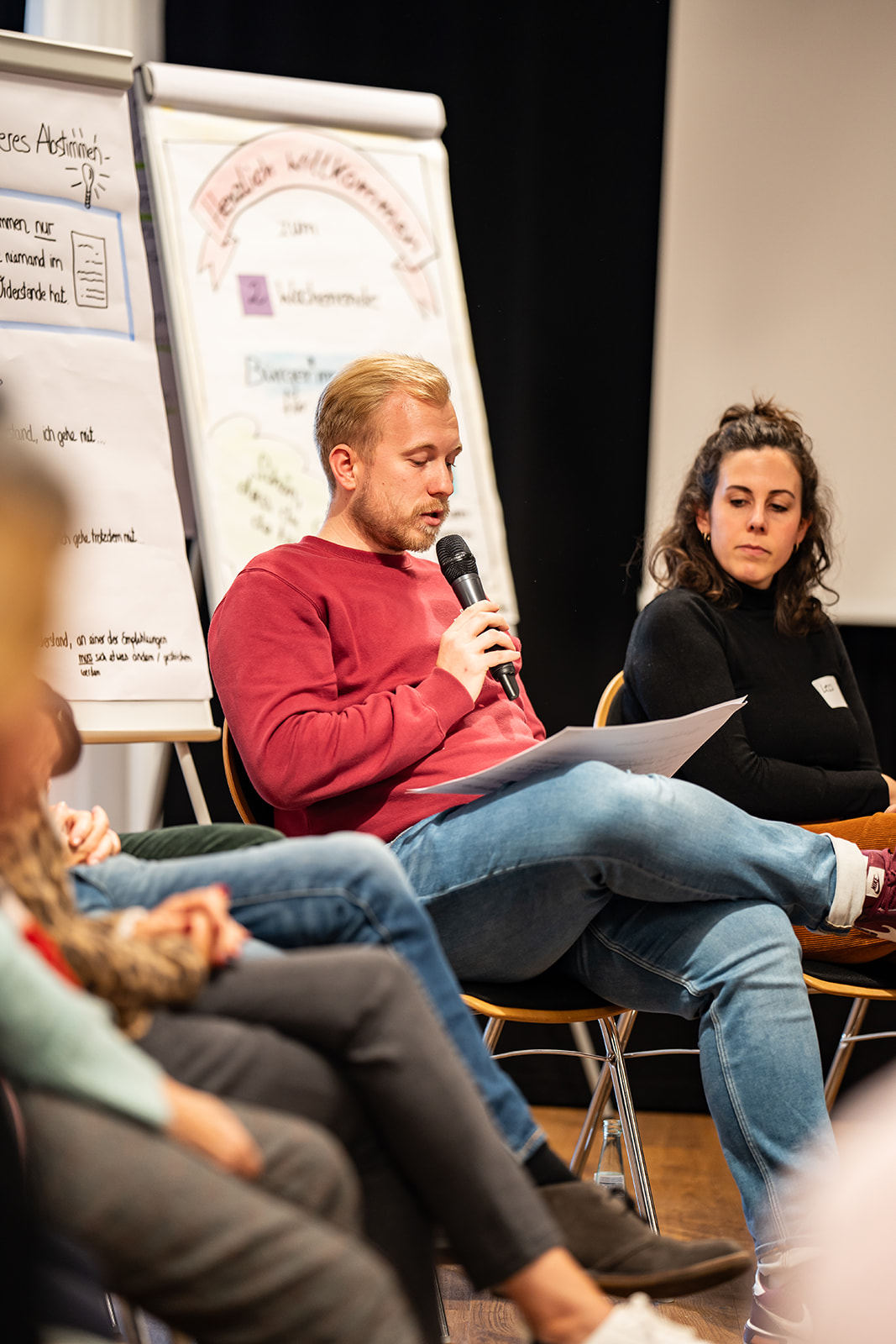 Ein Teilnehmer liest von einem Blatt Papier ab und spricht in ein Mikrofon. Er sitzt vor Flipcharts mit Ideen und Empfehlungen